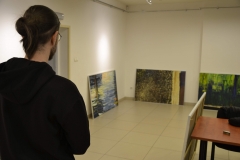 Po lewej stojący tyłem do obiektywu Marcin Derda w ciemnej bluzie. W głębi kadru oparte o ścianę trzy prace. Po prawej fragment stolika.