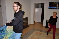 Po lewej Marcin Derda niosący jeden z obrazów. Po prawej  uśmiechnięta Krystyna Mikołajczak z CKiS w Koninie. W głębi wnęka drzwiowa i wiszący na ścianie ekran telewizora.