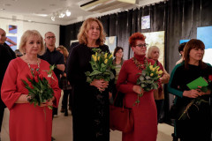Uczestnicy wernisażu. Na pierwszym planie stojące w rzędzie kobiety z bukietami kwiatów.