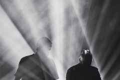 Dwóch muzyków przy konsolach pośród jasnych reflektorów. Zdjęcie czarno-białe.