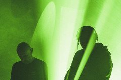 Dwóch muzyków przy konsolach. Jeden z nich ma słuchawki na uszach. Zielone światła reflektorów.