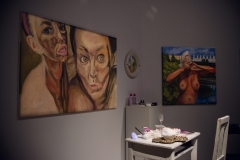 Dwa obrazy zawieszone na ścianie.  Obraz po lewej przedstawia twarze dwóch kobiet. Po prawej nagą kobietę widzianą od pasa w górę. Między nimi stoją biały stolik i krzesło. Na blacie leżą naczynia.