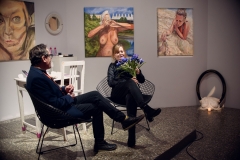 Robert Brzęcki i Ewa Gaj siedzą w fotelach na podwyższeniu podłogi. Kobieta trzyma w dłoniach granatowe kwiaty.