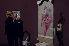 Uczestniczki wystawy oglądają obraz namalowany na szarym papierze. Przedstawia kobietę o różowej skórze, która w lewej dłoni trzyma telefon.