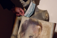 Szare popiersie manekina bez ramion. Z jego ucha zwisa niebieska maseczka. Przed nim leży obraz. Widziany fragment przedstawia twarz malarki.