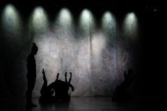 Tło sceny oświetlone światłem reflektorów. Po lewo ciemna sylwetka tancerza z kitką, obok niego na podłodze plątanina z rąk i nóg.