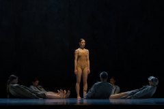W środku kadru tancerka ubrana w cieliste body. Wokół niej leżący na scenie tancerze zwróceni do niej stopami.