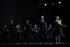 Zdjęcie grupowe. Tancerze ubrani w ciemne prochowce stoją na scenie. Wyciągają przed siebie ręce.