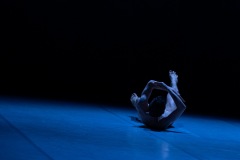 Tancerz leży na scenie. Z rąk stworzył obręcz, wyciąga stopy. Scena oświetlona błękitnym światłem, tło tonie w mroku.