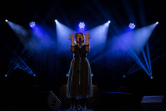 Dziewczyna w ciemnej tiulowej sukience przy mikrofonie na statywie. Na wysokości twarzy dwie dłonie. W tle światła reflektorów.
