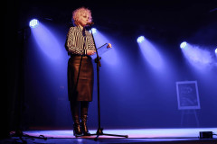 Na scenie Paulina Pachulska-Wojdak przy mikrofonie.  Za nią światła z czterech reflektorów.