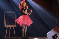 Na scenie dziewczynka w różowej tiulowej spódniczce, czarnej koszulce, z różową kokardą we włosach. W lewej dłoni trzyma mikrofon. Za nią snopy iskier. W głębi kadru na sztaludze plakat Festiwal Piosenki.