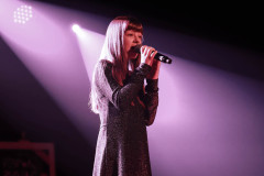 Na scenie długowłosa dziewczyna w brokatowej sukience. W obu dłoniach trzyma mikrofon blisko ust. Za  nią światła reflektorów. Zdjęcie w planie amerykańskim.