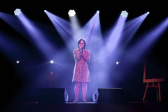Dziewczyna w sukience w grochy na scenie w planie ogólnym. Przed nią leżą dwa głośniki. Za nią krzyżujące się światła reflektorów.