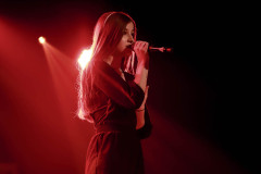Na scenie długowłosa dziewczyna w sukience. W prawej dłoni trzyma mikrofon. Za nią czerwona poświata. Zdjęcie w planie amerykańskim.
