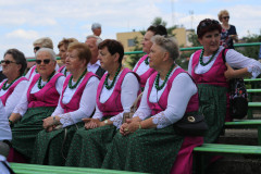 Członkinie zespołu ludowego na ławkach amfiteatru. Białe bluzki, różowe gorsety i spódnice, zielone zapaski.