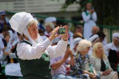 Kobieta w stroju ludowym robi zdjęcie telefonem. Ubrana w białą koszulę, zielony gorset i biały wysoki czepiec.