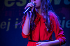 Dziewczynka z rozpuszczonymi długimi ciemnymi włosami, ubrana w czerwoną plisowaną sukienkę. W prawej ręce trzyma mikrofon.