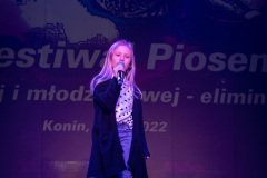 Na scenie blondwłosa dziewczynka w ciemnej narzutce, koszuli w groszki i niebieskich dżinsach.