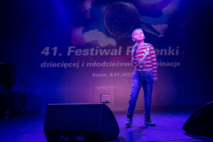 Na scenie w planie pełnym chłopiec z mikrofonem. Ubrany w bluzę w czerwono-białe pasy z reniferem i niebieskie dżinsy.