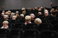 Seniorzy siedzący w ciemnych fotelach sali widowiskowej. Zdjęcie wykonane z dolnych rzędów.