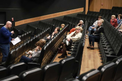 Seniorzy siedzący w fotelach kinowych, a przed nimi mężczyzna w granatowym swetrze z podkładką z kartką. W prawej dłoni trzyma mikrofon. Zdjęcie wykonane zza szóstego rzędu.