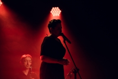 Zdjęcie utrzymane w czerwonej i czarnej tonacji. Ciemna sylwetka kobiety przy mikrofonie, za nią muzyk.