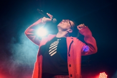 Kobieta w czewronym płaszczu odchyla do tyłu głowę. W prawej ręce trzyma przybliżony do ust mikrofon, lewą zgiętą w łokciu unosi do góry. osi
