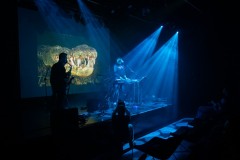 Mroczne zdjęcie. Snopy światla na scenie, dwóch muzyków, a za nimi zdjęcie paszczy krokodyla.