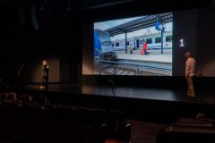 Jurorzy na scenie zwróceni w stronę ekranu, na którym wyświetlona jest fotoggrafia FotoBitwy, opatrzona numerem 1.  Zdjęcie przedstawia dworzec kolejowy, pociągi i stojące na peronie dwie osoby.