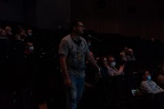 Mężczyzna w dżinsach, szarej koszulce i okularach stoi przy mikrofonie. Za nim siedzą uczestnicy FotoBitwy.