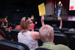Kobieta odwrócona plecami do obiektywu, widziana z prawego profilu. Unosi w górę żółtą kartkę. Obok niej siedzi osoba. Przed nią rzędy foteli i rozmyta osoba na scenie w głębi kadru.