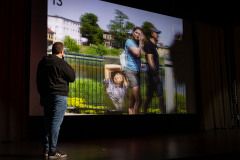 Mężczyzna na scenie stoi tyłem do obiektywu, trzymając mikrofon. Komentuje zdjęcie opatrzone numerem 13.