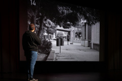 Mężczyzna stoi na scenie tyłem do obiektywu. Trzyma mikrofon i komentuje czarno-białe zdjęcie opatrzone numerem 14. Zdjęcie przedstawia chodnik przed dawnym DT Centrum.