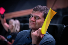 Zdjęcie portretowe. Uśmiechnięty mężczyzna patrzy w obiektyw. Przy twarzy trzyma żółtą kartkę.