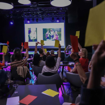 Uczestnicy siedzący tyłem do obiektywu. Podnoszą w górę żółte i czerwone kartki. Przed nimi ekran i wyświetlony slad z dowma zdjęciami.