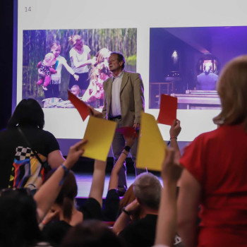 Na pierwszym planie stojąca tyłem do obiektywu kobieta w czerwonej koszulce. Obok dłonie trzymające żółte i czerwone kartki. Na scenie Mirosław Jurgielewicz. Za nim wyświetlony slajd z dwoma zdjęciami.