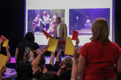 Na pierwszym planie stojąca tyłem do obiektywu kobieta w czerwonej koszulce. Obok dłonie trzymające żółte i czerwone kartki. Na scenie Mirosław Jurgielewicz. Za nim wyświetlony slajd z dwoma zdjęciami.
