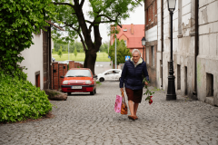 Idąca ulicą Śliską w Koninie starsza kobieta. W prawej ręce trzyma torby, w lewej - czerwoną różę. W tle dwa auta: czerwone i białe.(zdjęcie nr 12)