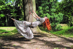 Rudowłosa kobieta w długiej sukni w kwiatowy deseń wygina się na huśtawce zawieszonej na drzewie. Autor: Jan Witkowski.