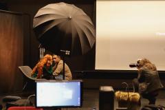 Sesja zdjęciowa. Rudowłosa modelka w fotelu, obok niej kucający Patryk Koszela, przed ną fotografującą. Na pierwszym planie rozświetlony monitor i rozłożony parasol fotograficzny.
