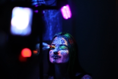 Młoda długowłosa kobieta podczas malowania farbami UV. Na pierwszym planie zamglony szkielet lampy.