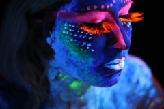 Kobieta pomalowana farbami UV w kolorach niebieskim, różowym i pomarańczowym. Za zamknięte powieki zakończone długimi rzęsami.