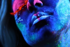 Wizaż farb UV. Niebieska kobieca twarz z różowymi nosem i powiekami oraz długimi, pomarańczowymi rzęsami.