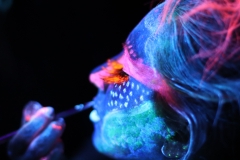 Profil kobiety podczas malowania farbami UV. Jej twarz pokrywają kolory niebieski, różowy i pomarańczowy.