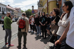 Uczestnicy spaceru stoją na chodniku wzdłuż żóltego budynku przy ulicy 3 Maja. W głębi szyld sklepu Rossmann, latarnia ze znakiem zakazu zatrzymywania się.