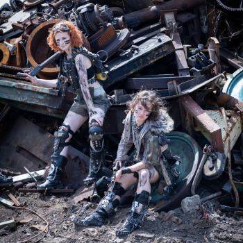 Dwie modelki w strojach w stylu Mad Max. Pozują na tle piętrzącej się sterty złomu. Jedna z nich stoi, druga siedzi.