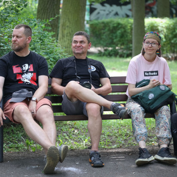 Na ławce w parku siedzą mężczyźni w ciemnych koszulkach i spodenkach do kolan oraz kobieta w chustce na głowie, różowej koszulce i wzorzystych spodniach. Obok ławki ciemny plecak.