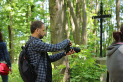 Mężczyzna sfotografowany z prawego profilu, W dłoniach trzyma aparat i robi zdjęcie patrząc w wyświetlacz. Ubrany w koszule w kartę. Na plecach czarny plecak. W głębi drzewa i krzyż.