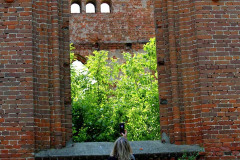 Odwrócona plecami do obiektywu kobieta stoi przed ostrołukowym oknem zrujnowanego kościoła z czerwonej cegły.